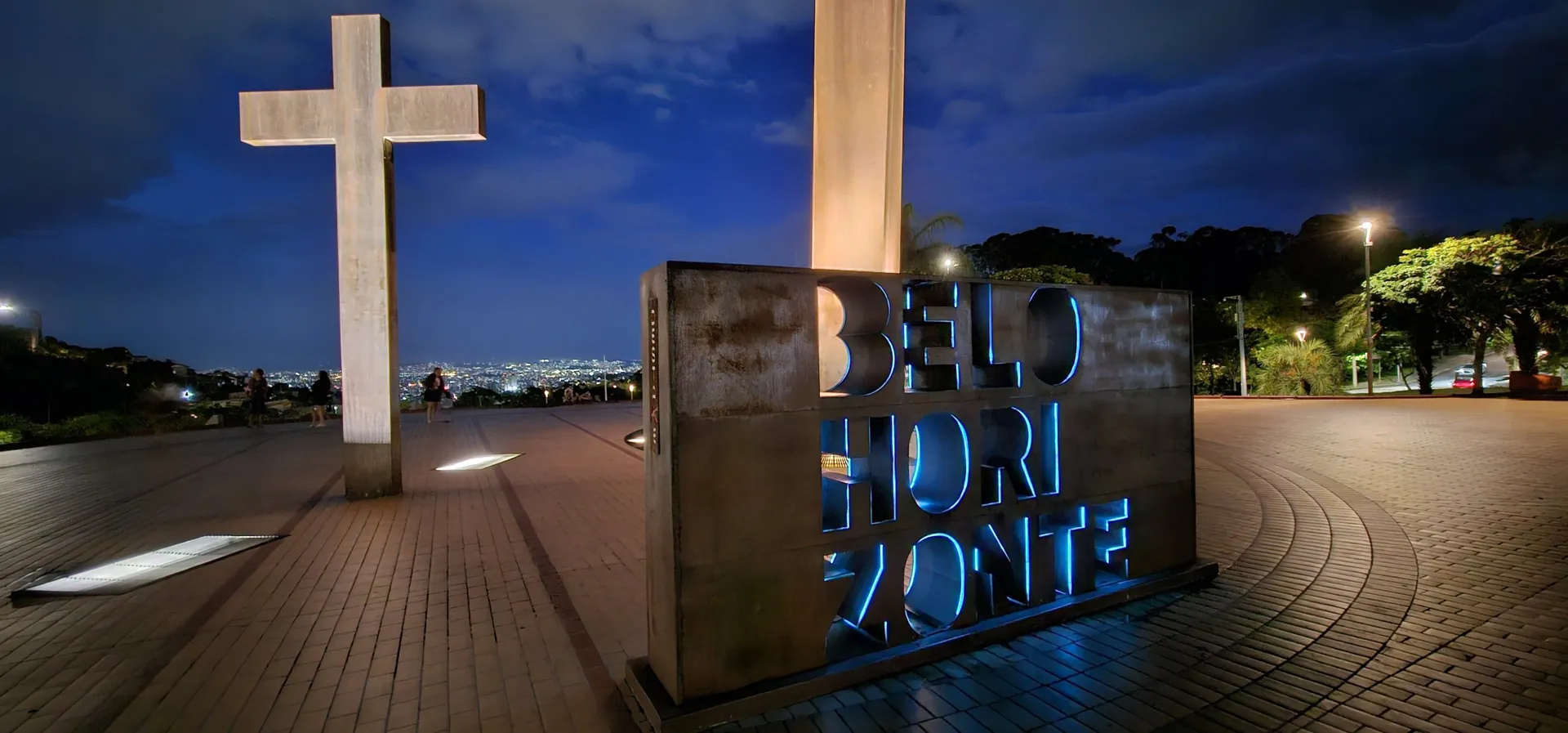 Letreiro de Belo Horizonte iluminado em azul à noite na Praça do Papa, com a cruz e as luzes da cidade ao fundo