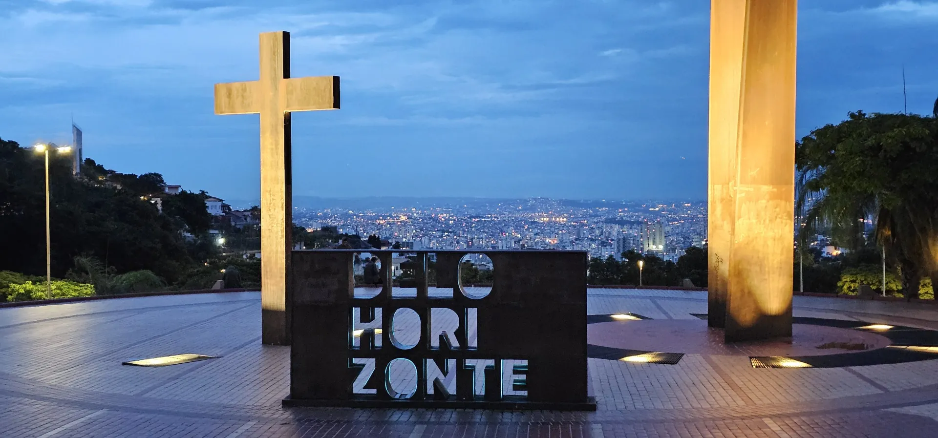 Cruz e letreiro de Belo Horizonte na Praça do Papa, no Mangabeiras, com a cidade iluminada ao fundo no fim da tarde