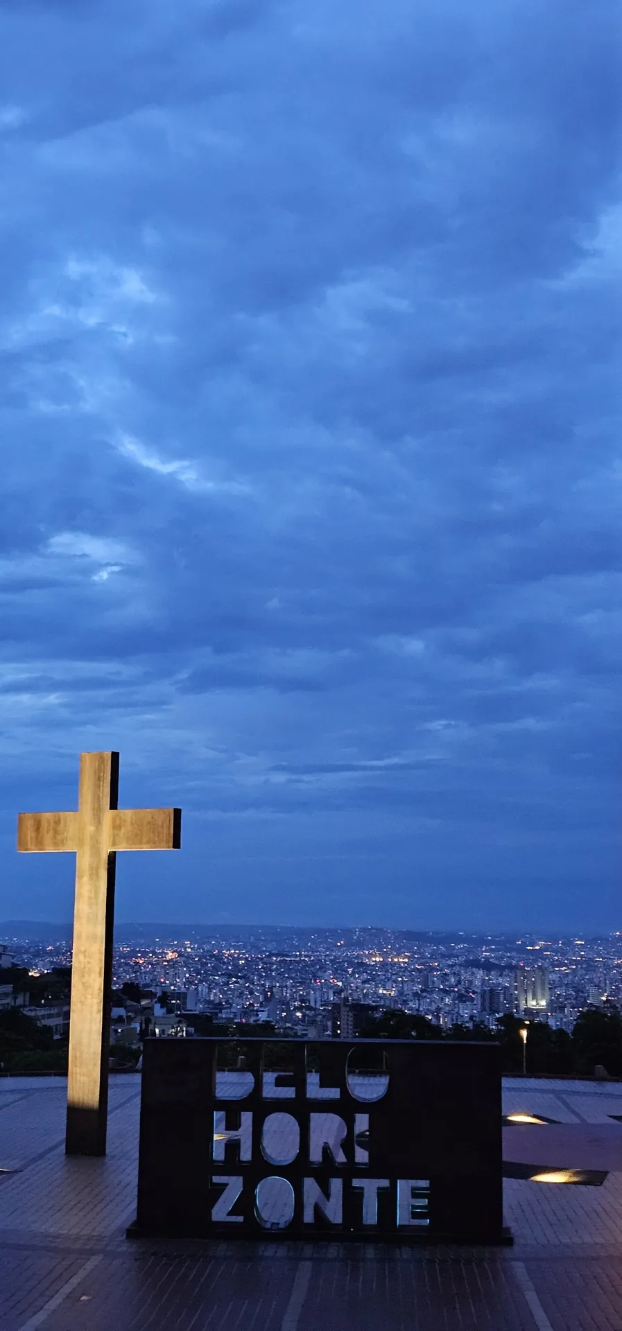 Cruz e letreiro de Belo Horizonte na Praça do Papa com o céu azul e a vista da cidade iluminada ao anoitecer
